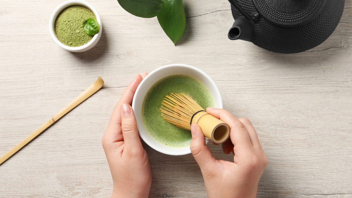 hands holding a whisk preparing matcha tea in a lowl. On the table there is a iron tea pot and a bowl of matcha powder