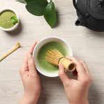 hands holding a whisk preparing matcha tea in a lowl. On the table there is a iron tea pot and a bowl of matcha powder