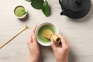 hands holding a whisk preparing matcha tea in a lowl. On the table there is a iron tea pot and a bowl of matcha powder