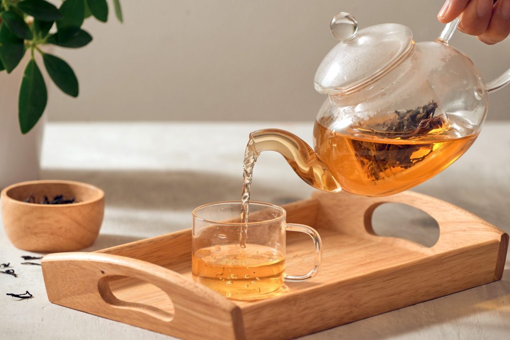 a hand pouring tea from a glass tea pot into a glass cup on a wooden tray.