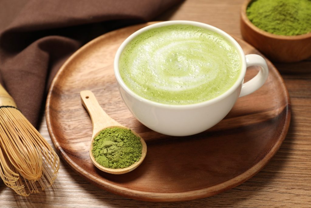 A matcha latte in a white cup on a wooden tray. Next to the cup is a wooden spoon with matcha powder in it and a bamboo whisk.