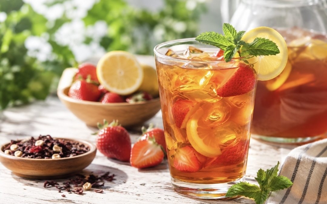 A glass of refreshing iced tea with ice cubes and fruit sits on a table with a bowl of strawberries and sliced lemon in the background.